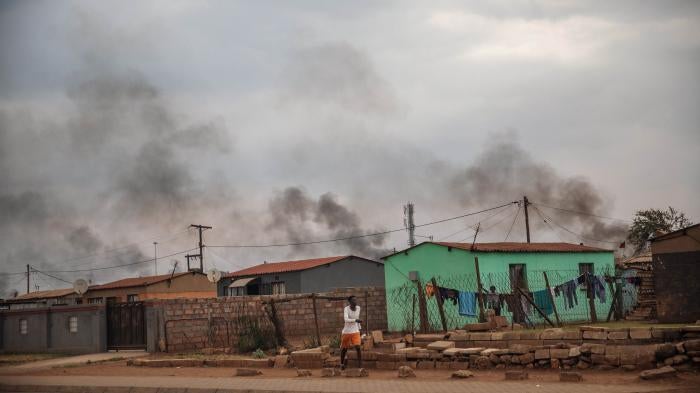 A man walks in front of a buildings, with smoke rising in the background
