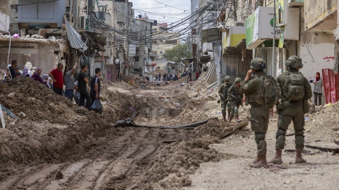 Israeli soldiers look on as Palestinian men and women carry their belongings on May 2, 2025 amid ongoing Israeli military operations and demolition of homes in Tulkarem refugee camp. Israeli forces had emptied the refugee camp of its inhabitants weeks prior.