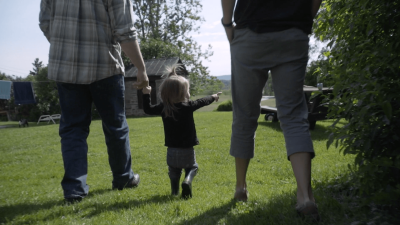 A young child stands between her parents in their garden, pointing into the distance